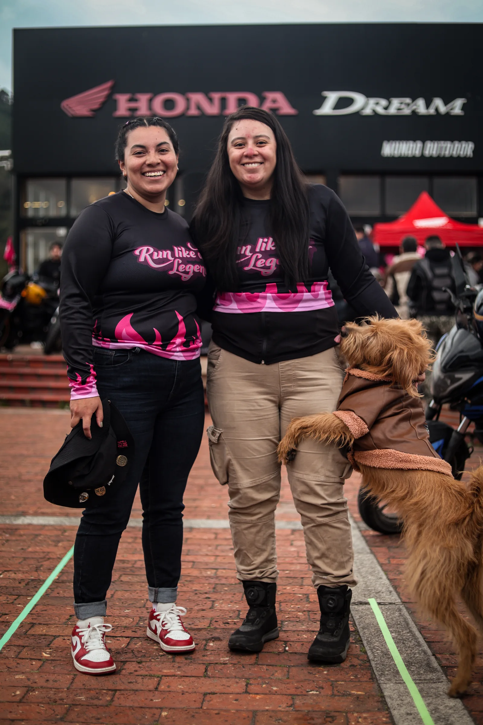 Dos participantes sonríen junto a un perro frente al letrero de Honda Dream.